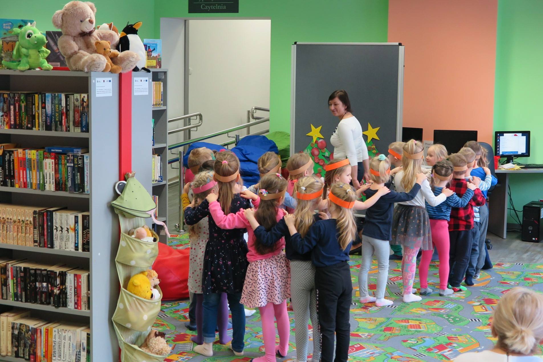 Wnętrze biblioteki. Razem z bibliotekarką Anią Wiśniewską dzieci bawią się, w kolorowych opaskach ustawiają się jeden za drugim tworząc mały pociąg.