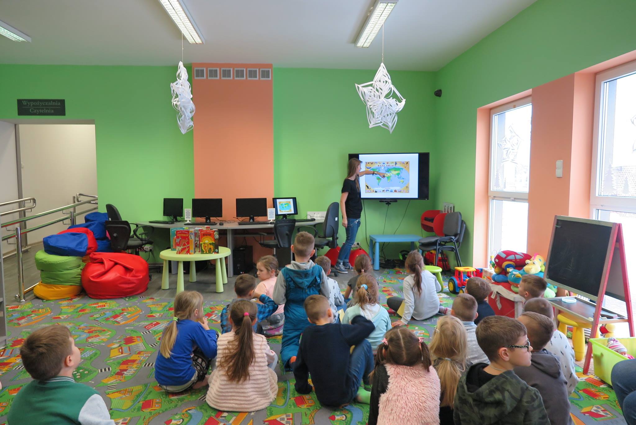 Wnętrze biblioteki. Na pufach siedzą dzieci i oglądają na monitorze prezentację dotyczącą kontynentów. Lekcję biblioteczną prowadzi Aleksandra Cybulska.
