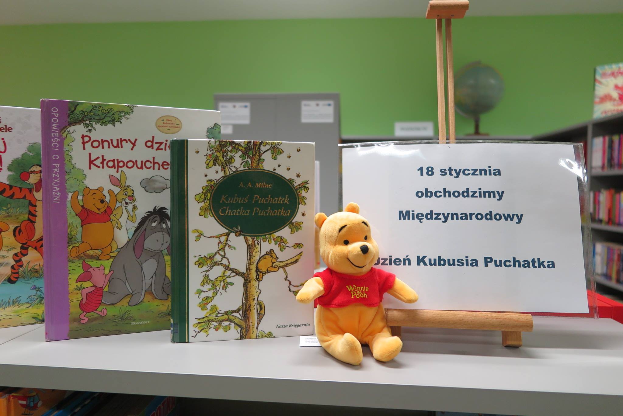 Na półce mini sztaluga z plakatem 18-stycznia obchodzimy Międzynarodowy Dzień Kubusia Puchatka obok książki Kubusia Puchatka oraz pluszowa maskotka Kubuś Puchatek.