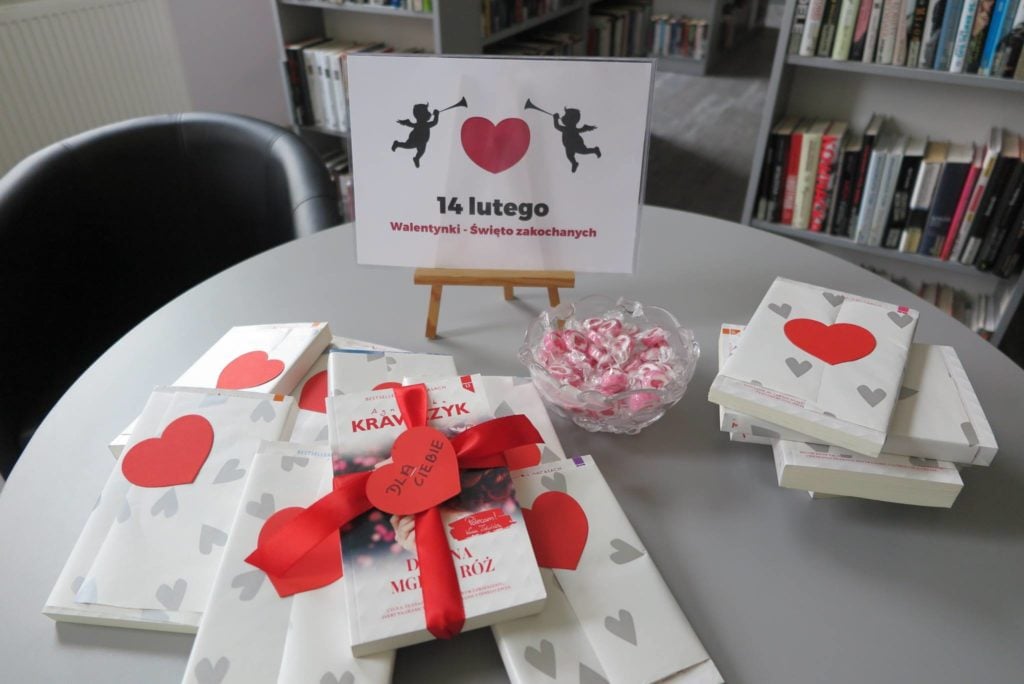 Na stoliku mała sztaluga z plakatem 14 luty - Walentynki- święto zakochanych. Obok książki owinięte w papier  w serduszka i miseczka z cukierkami.
