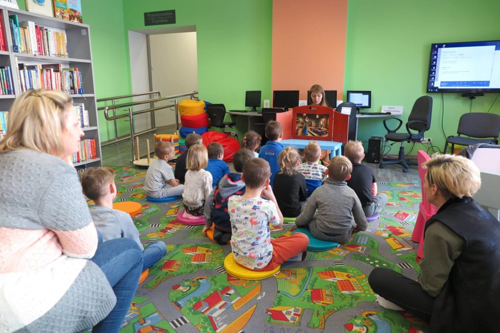 Wnętrze biblioteki. Na pufach siedzą dzieci z grupy V z Przedszkola nr 3 słuchają teatrzyku Kamishibai "Królewna Śnieżka i siedmiu krasnoludków" ,czyta bibliotekarka Ola Cybulska.