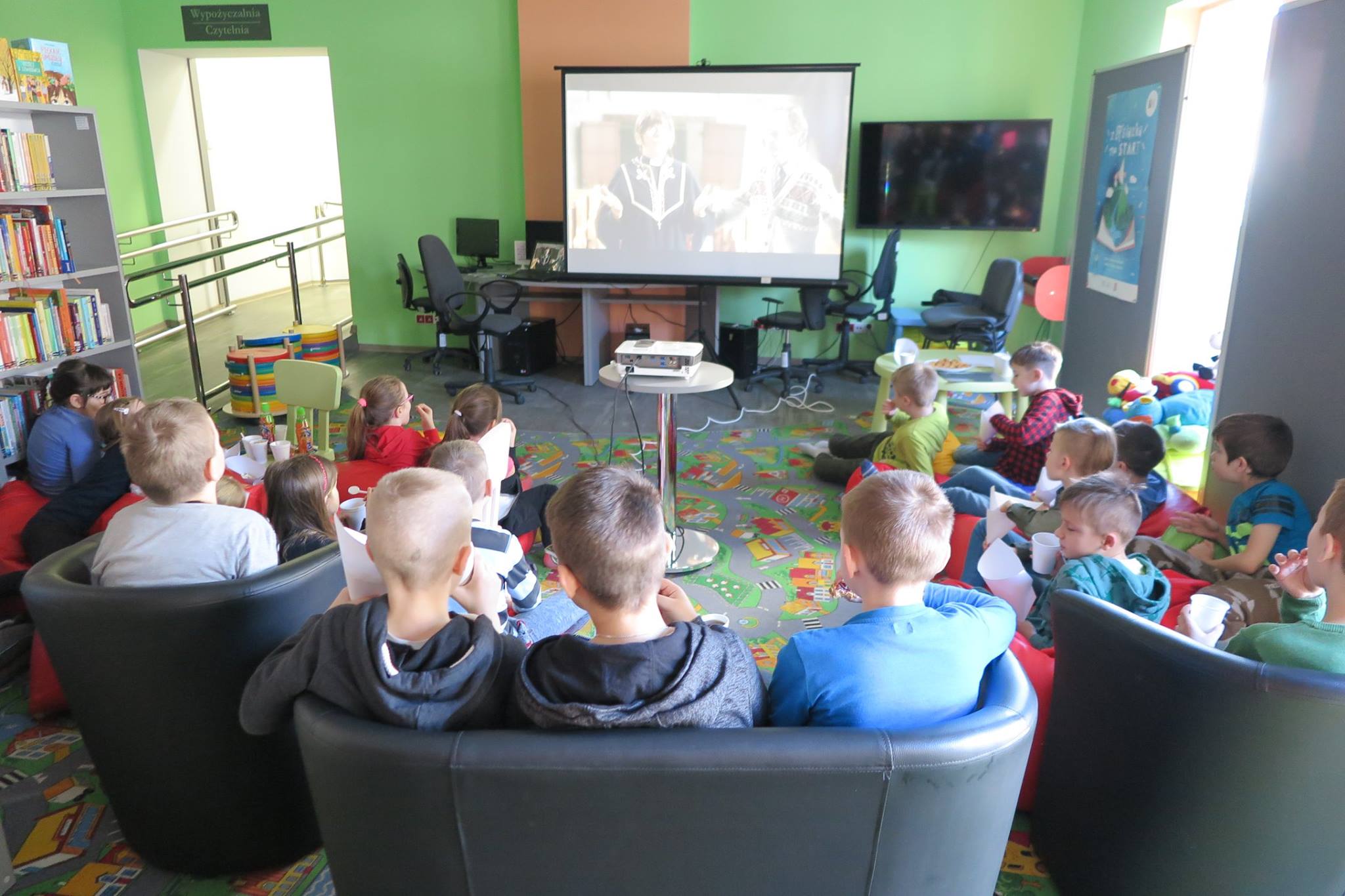 Wnętrze biblioteki. Na kanapach siedzą dzieci z DKK  i oglądają  film na podstawie książki, którą omawiali w  ubiegłym miesiącu. W tle rzutnik i monitor na którym wyświetlany jest film. 