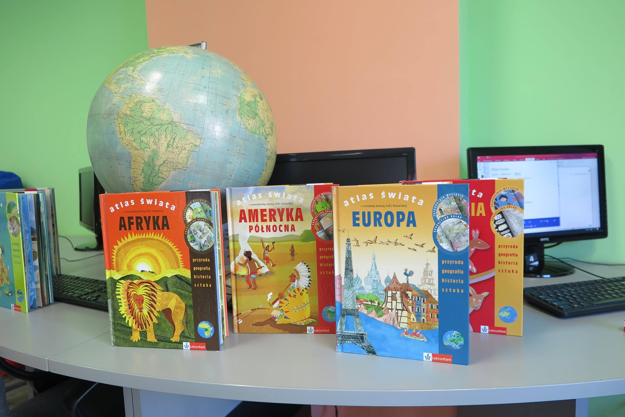 Na stoliku komputery, globus i pięć atlasów świata z kontynentów takich jak Afryka, Ameryka Północna, Europa.