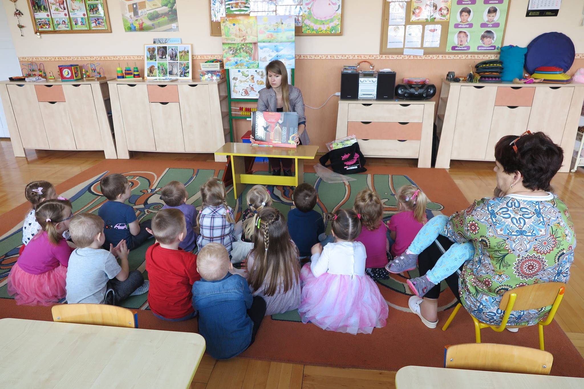Wnętrze przedszkola nr 1, na kolorowej wykładzinie siedzą dzieci i słuchają teatrzyku  "Słowik",który czyta bibliotekarka Ola Cybulska.