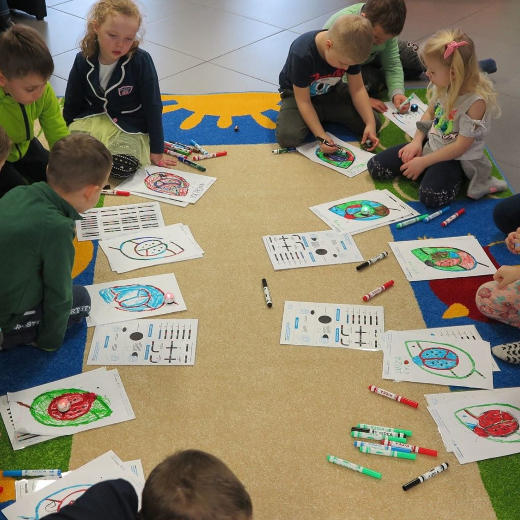 Na kolorowej wykładzinie siedzą dzieci i biorą udział w warsztatach z kodowania ozobotów, Każdy z uczestników rysuje trasę dla ozobotów.