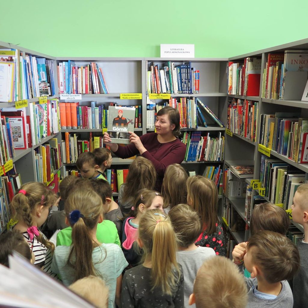 Wnętrze biblioteki. Bibliotekarka Ania Wiśniewska między regałami książek pokazuje dzieciom książkę i opowiada jak powstają książki.