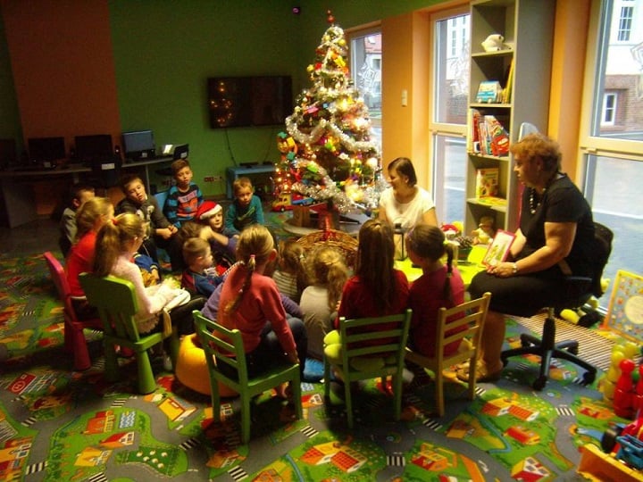 Na krzesełkach siedzą dzieci obok dwie biblioteakarki wspólnie spędzają świąteczny czas na środku choinka świąteczna.