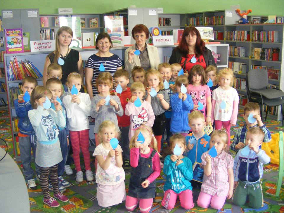 Wnętrze biblioteki. Grupa dzieci z opiekunkami i bibliotekarkami trzyma w dłoni wykonaną z papieru niebieską kroplę wody.