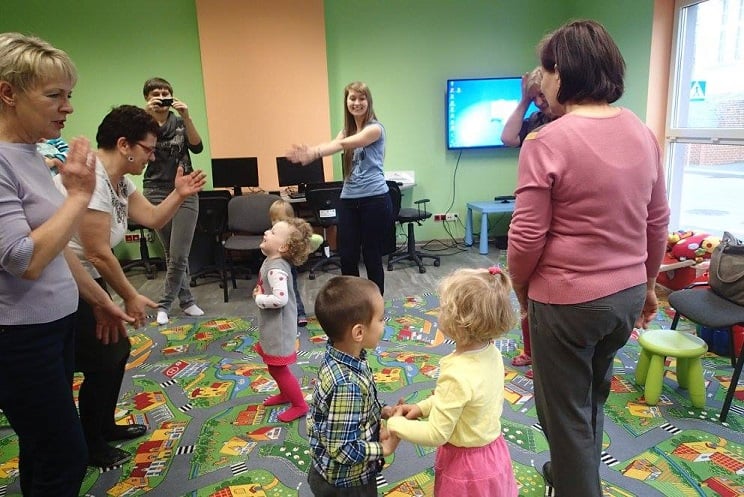 Na kolorowej wykładzinie bawią się dzieci z opiekunami i bibliotekarką Aleksandrą.