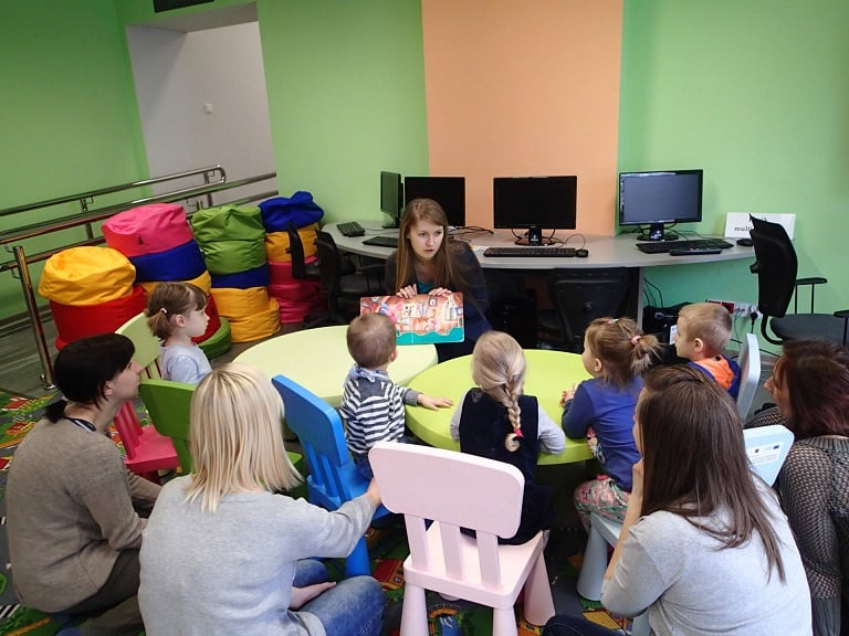 Przy stolikach siedzą dzieci, bibliotekarka Aleksandra Cybulska czyta książeczkę "Wielkie sprzątanie" z Kubusiem Puchatkiem.  
