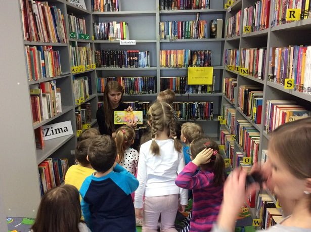 Między regałami z książkami stoi grupa dzieci z Przedszkola '' U misia'', bibliotekarka Aleksandra pokazuje bajki i opowiadania polskich autorów.