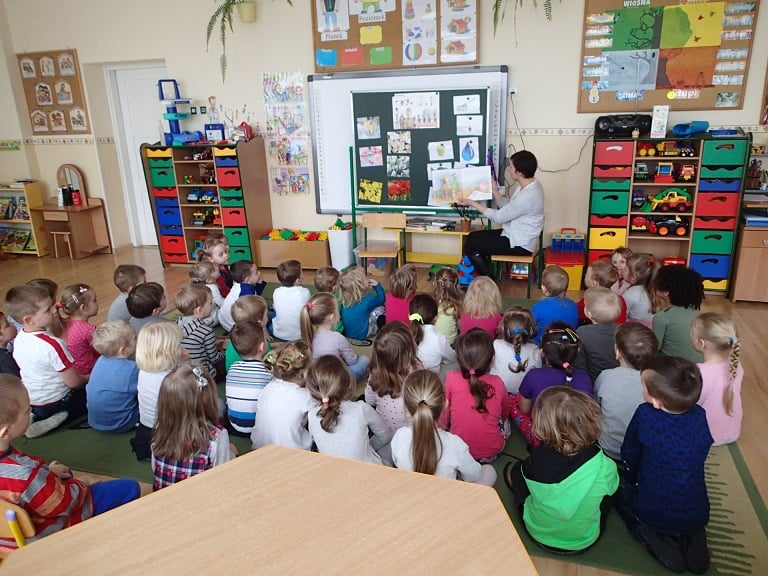 Wnętrze Przedszkola nr 1 , grupa dzieci siedzi na kolorowej wykładzinie, uważnie słucha jak czyta bibliotekarka Ania bajki o wiośnie i Tupciu Chrupciu. 