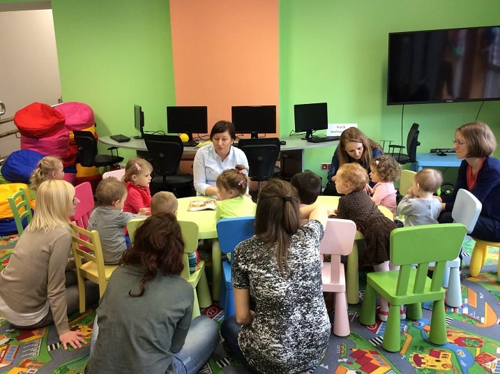 Przy stolikach siedzą dzieci z opiekunami, bibliotekarka Ania Wiśniewska czyta opowiadania z czasopisma o Kubusiu Puchatku, który bawił się z przyjaciółmi na huśtawce. W tle na ścianie telewizor.
