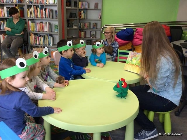 Przy zielonych stolikach siedzi sześcioro dzieci w zielonych opaskach na głowie, bibliotekarka Aleksandra Cybulska czyta  książkę "Żaba Ferdynand". 