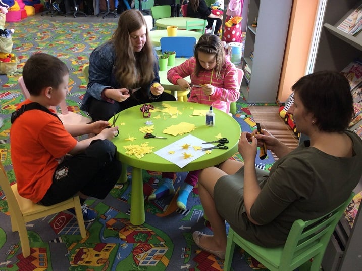 Przy stoliku siedzi trójka dzieci i bibliotekarka Ania Wiśniewska wspólnie wycinają z żółtego papieru żonkile. 