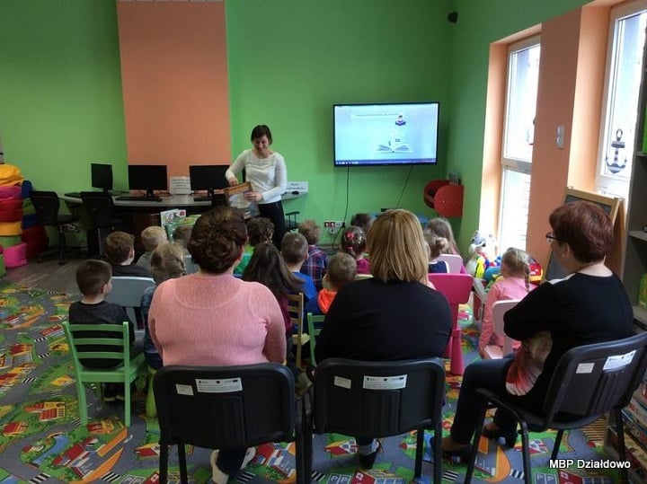 Na krzesełkach siedzą dzieci z opiekunami, lekcję prowadzi bibliotekarka Ania Wiśniewska na temat jak powstaje książka , która później trafia do biblioteki. 