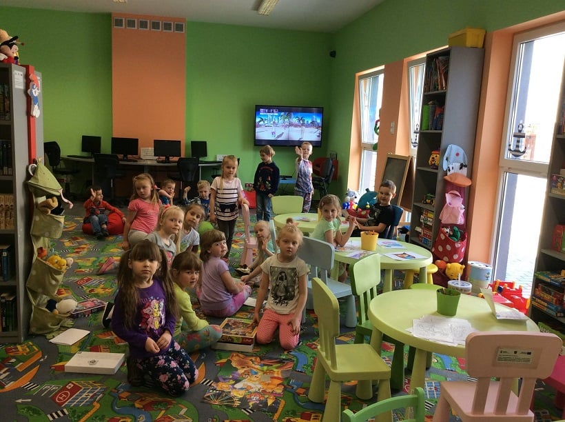 W oddziale dla dzieci w bibliotece bawią się przedszkolaki, w tle telewizor z prezentacją bajkową. 