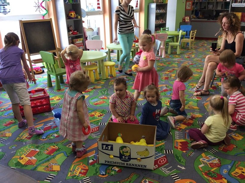 Wnętrze biblioteki. Na kolorowej wykładzinie bawi się grupa dzieci, uczą się segregować zabawki.