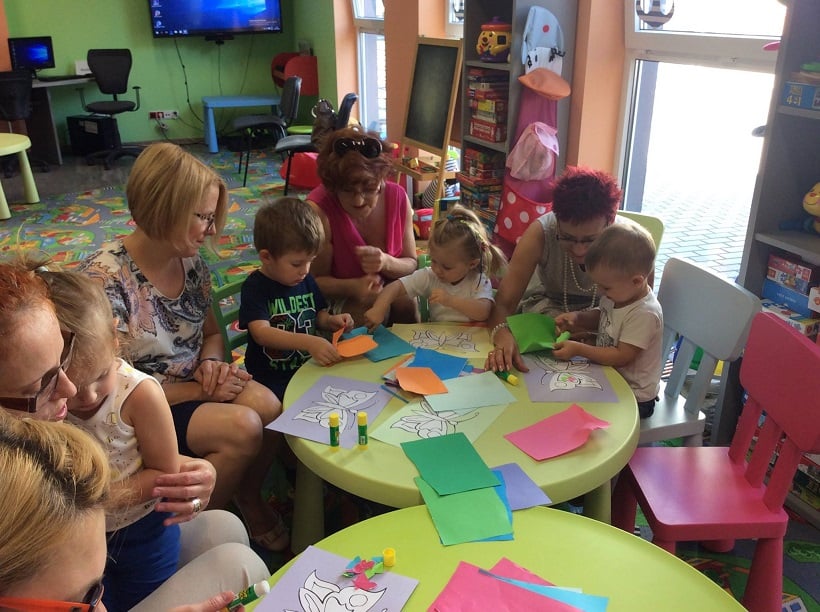 Przy stolikach siedzą dzieci z Klubu Malucha wspólnie z opiekunkami metodą wydzieranki kolorują motylki.