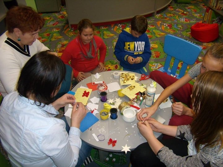 Przy stoliku siedzi grupa dzieci i dwie bibliotekarki wspólnie wykonują kartki świąteczne.