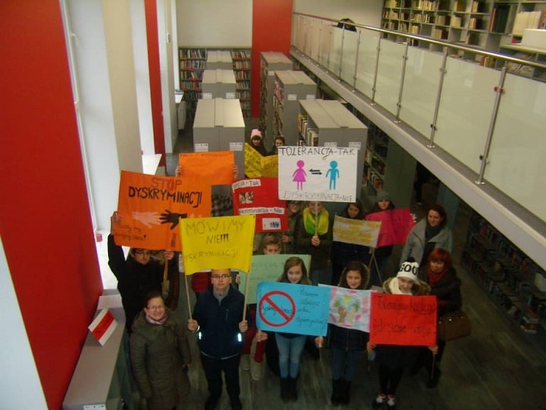Wnętrze biblioteki, grupa dzieci z transparentami na temat tolerancji oraz dyskryminacji - "Tolerancja - TAK, Dyskryminacja - NIE".