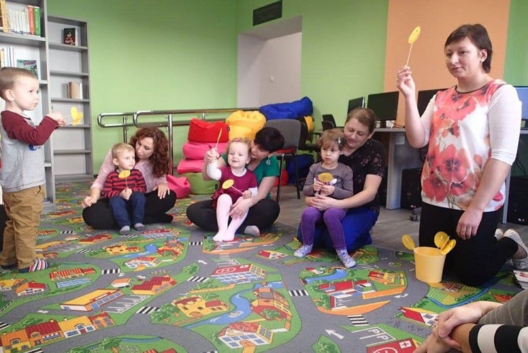Na kolorowej wykładzinie siedzą dzieci z opiekunkami wspólnie biorą udział w zabawie z bibliotekarką Anią Wiśniewską. 