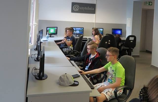 Wnętrze biblioteki, mediateka. Przy komputerach siedzą dzieci biorą udział w warsztatach z nauki programowania przy użyciu  Minecraft wersji edukacyjnej.