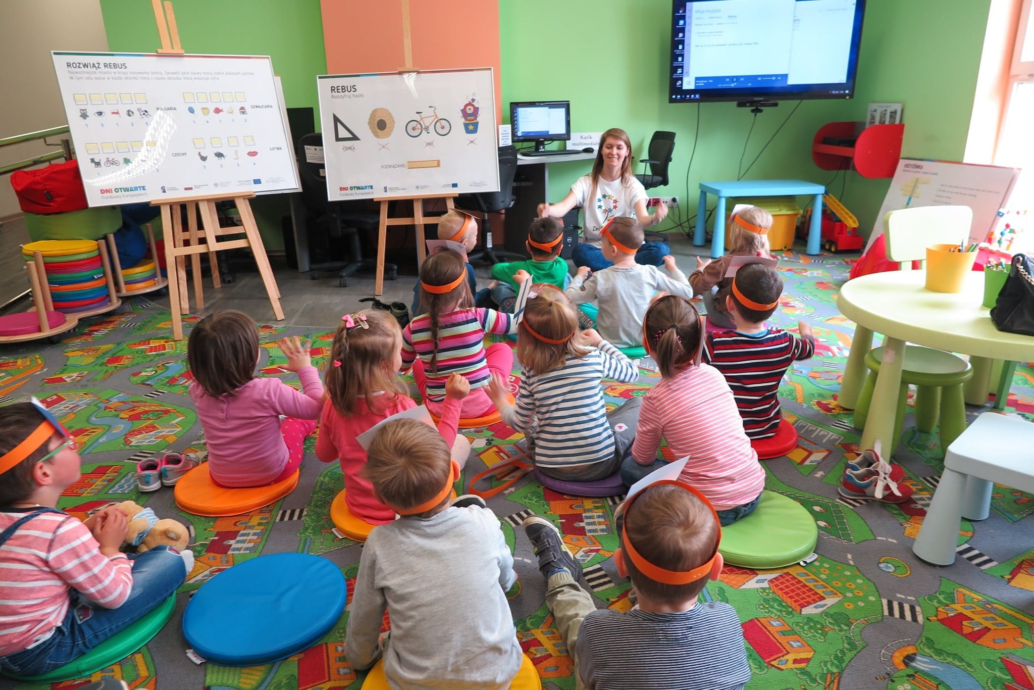 Wnętrze biblioteki. Na kolorowych pufach siedzą dzieci. Biorą udział w lekcji bibliotecznej edukacyjnej pt. "Poznajemy kraje Unii Europejskiej". Lekcję prowadzi bibliotekarka Ola Cybulska.