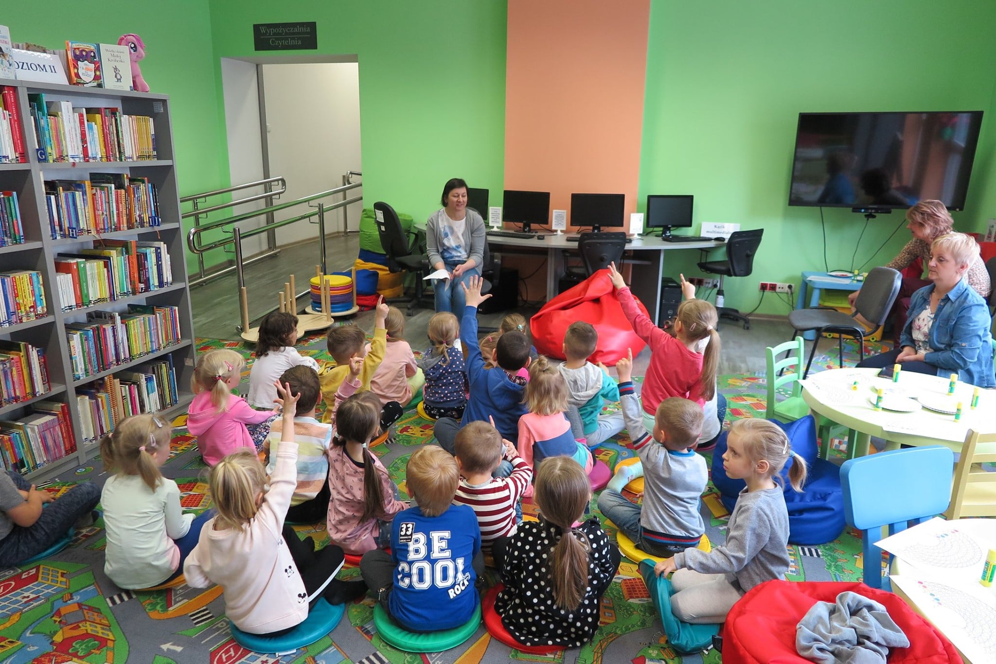 Wnętrze biblioteki. Dzieci siedzą na pufach i biorą udział w zajęciach o kolorach, prowadzi je bibliotekarka Ania Wiśniewska.