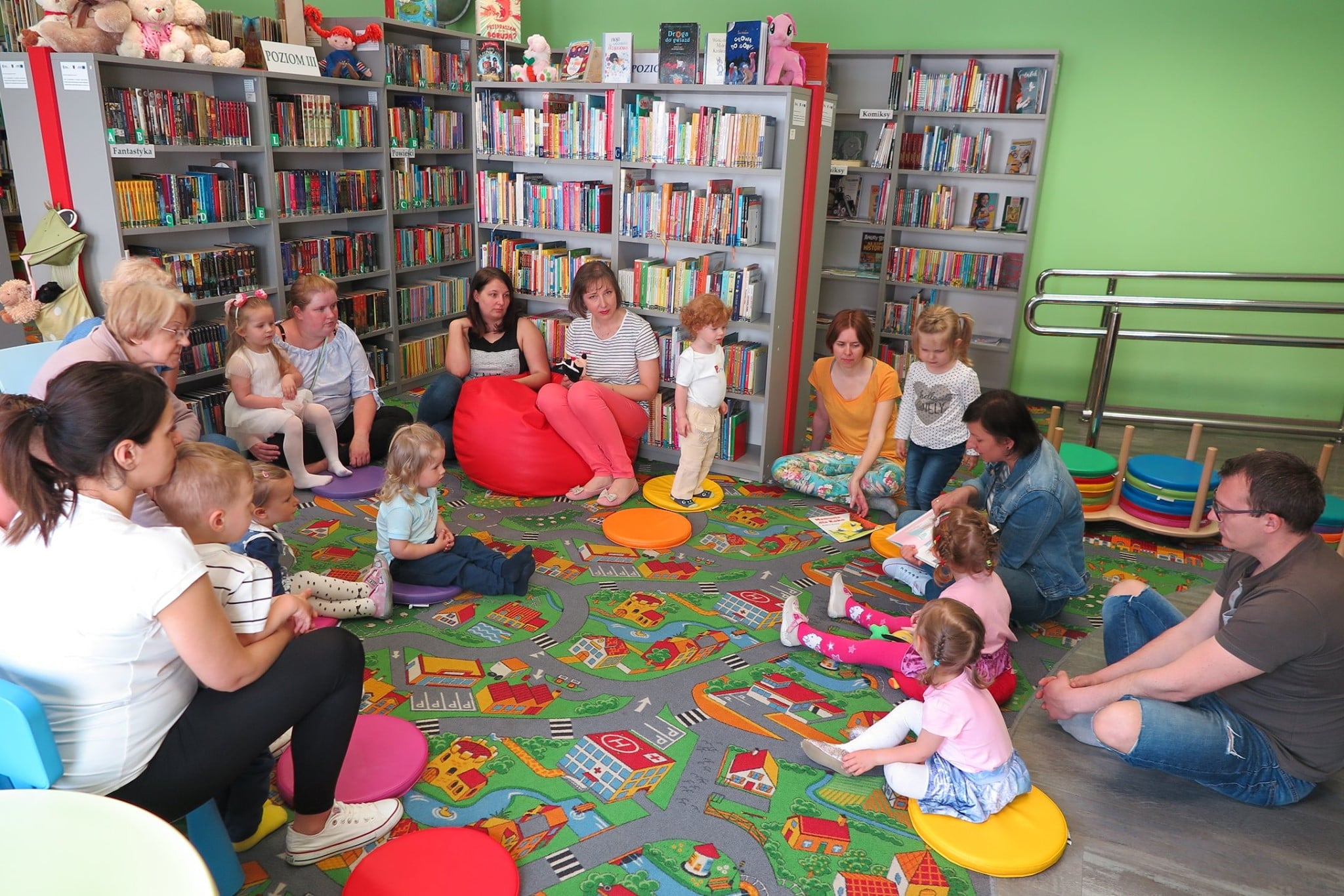 Wnętrze biblioteki. Na pufach siedzą dzieci z Klubu Malucha spotkały się z okazji dnia mamy. Książkę "Mama dla Koko". K.Kaszy czyta bibliotekarka Ania Wiśniewska.