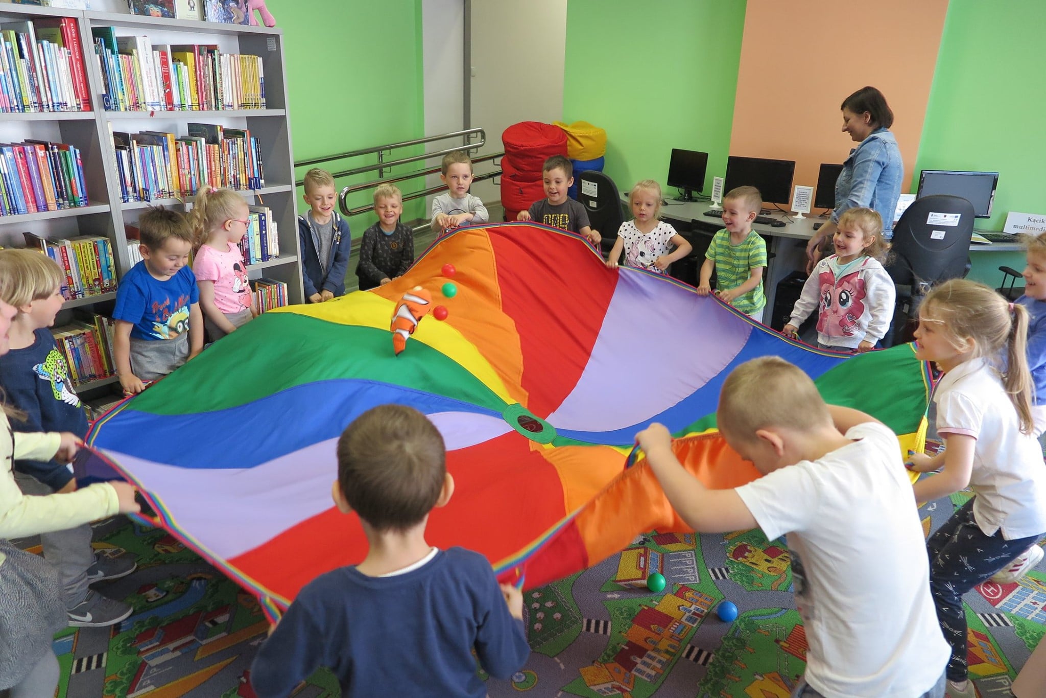 Wnętrze biblioteki. Dzieci trzymają kolorową chustę, obok bibliotekarka Ania Wiśniewska.