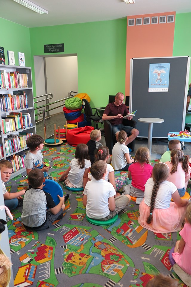 Wnętrze biblioteki. Na pufach siedzą dzieci i słuchają opowiadania pana Łukasza Schulza - absolwenta Wychowania Fizycznego.