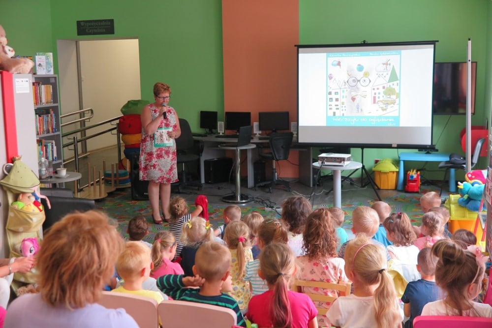 Wnętrze biblioteki. Spotkanie dzieci z Renatą Buczyńską autorką książki ,,Wierszyki do oglądania''. W tle prezentacja wiersza na monitorze.