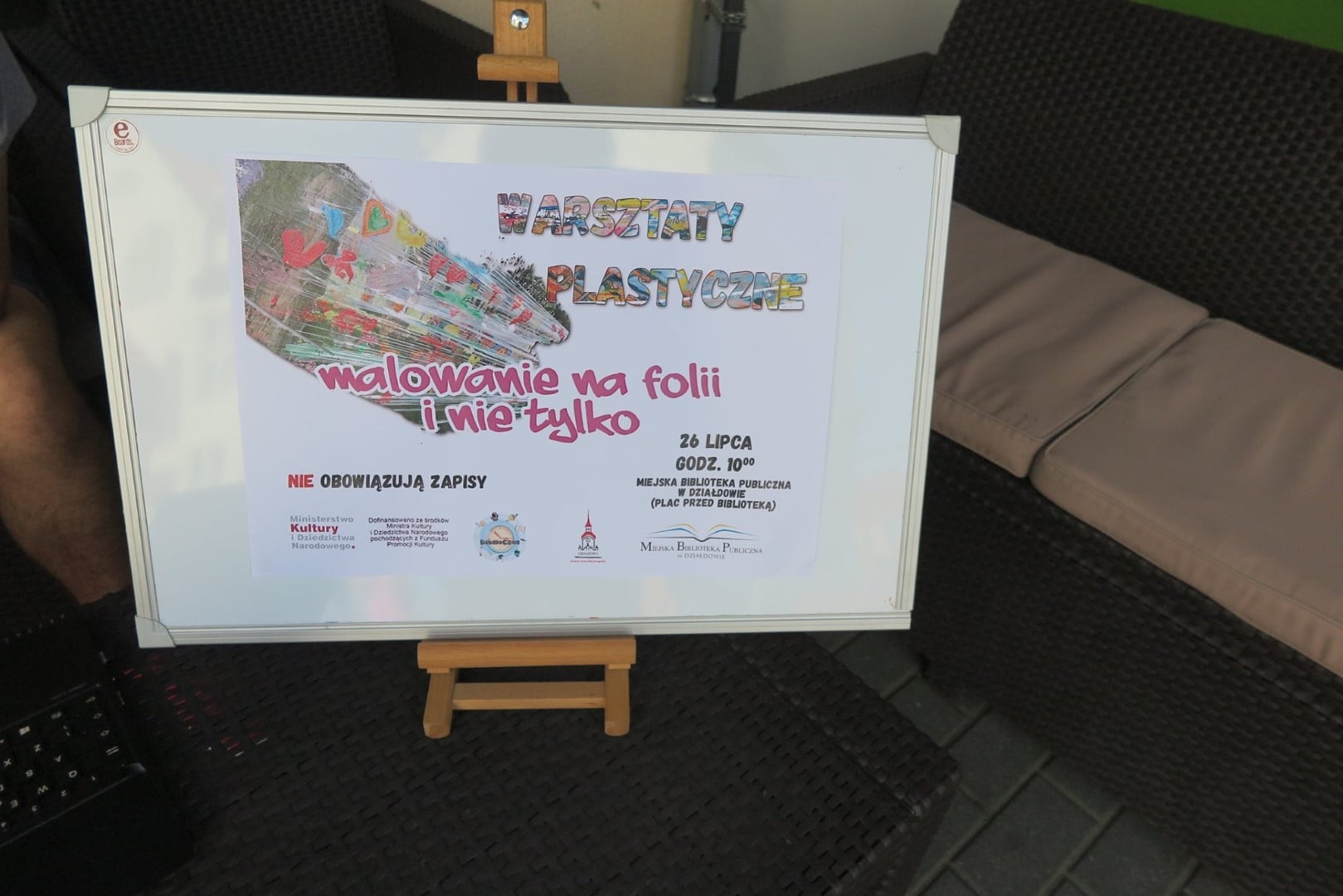 Na sztaludze plakat MBP ''Warsztaty Plastyczne''. Malowanie na folii i nie tylko. 26 lipca godz.10:00.  