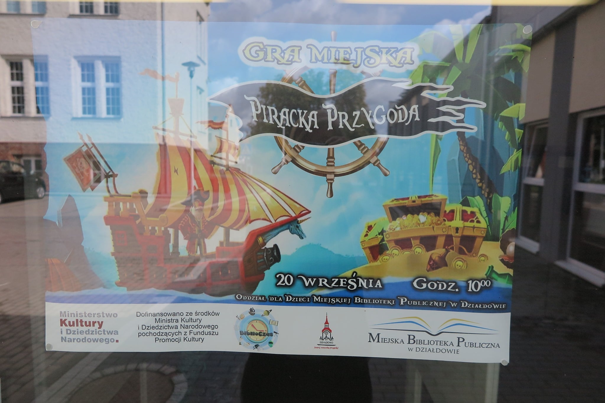 Na szybie plakat z napisem ''Gra Miejska Piracka Przygoda'', na plakacie piracki statek, i skarb piratów. 