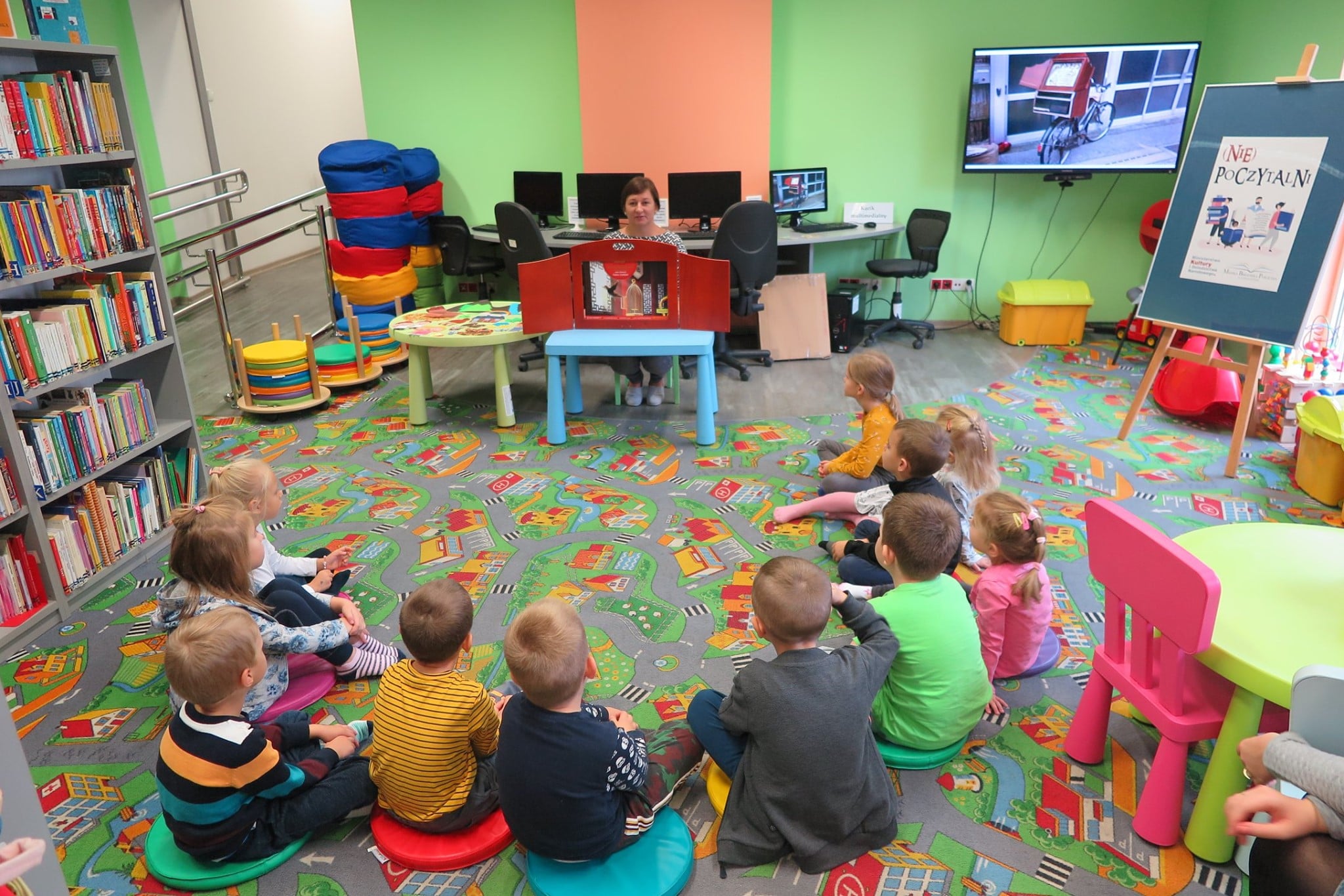 Wnętrze biblioteki ,oddział dla dzieci. Dzieci siedzą na pufach i słuchają teatrzyku kamishibai, który czyta bibliotekarka Ania Wiśniewska.  