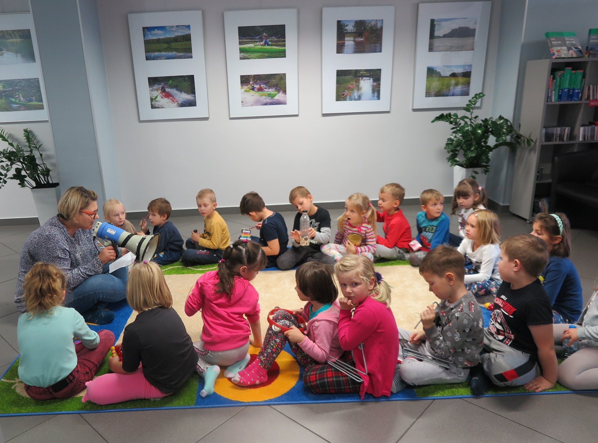 Wnętrze biblioteki,mediateka.Dzieci z przedszkola nr 3 grupa ''Motylki''  siedzą na kolorowym dywanie i biorą udział w warsztatach muzycznych. Zajęcia prowadzi bibliotekarka Dorota Kusik. 