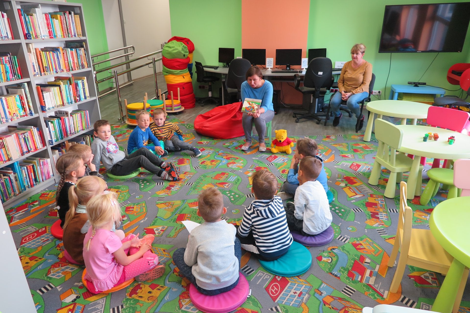 Wnętrze biblioteki, dzieci z Przedszkola nr 4  siedzą na pufach w kole i słuchają opowiadania o Kubusiu Puchatku i jego przyjaciołach ze Stumilowego Lasu. Zajęcia prowadzi Ania Wiśniewska.