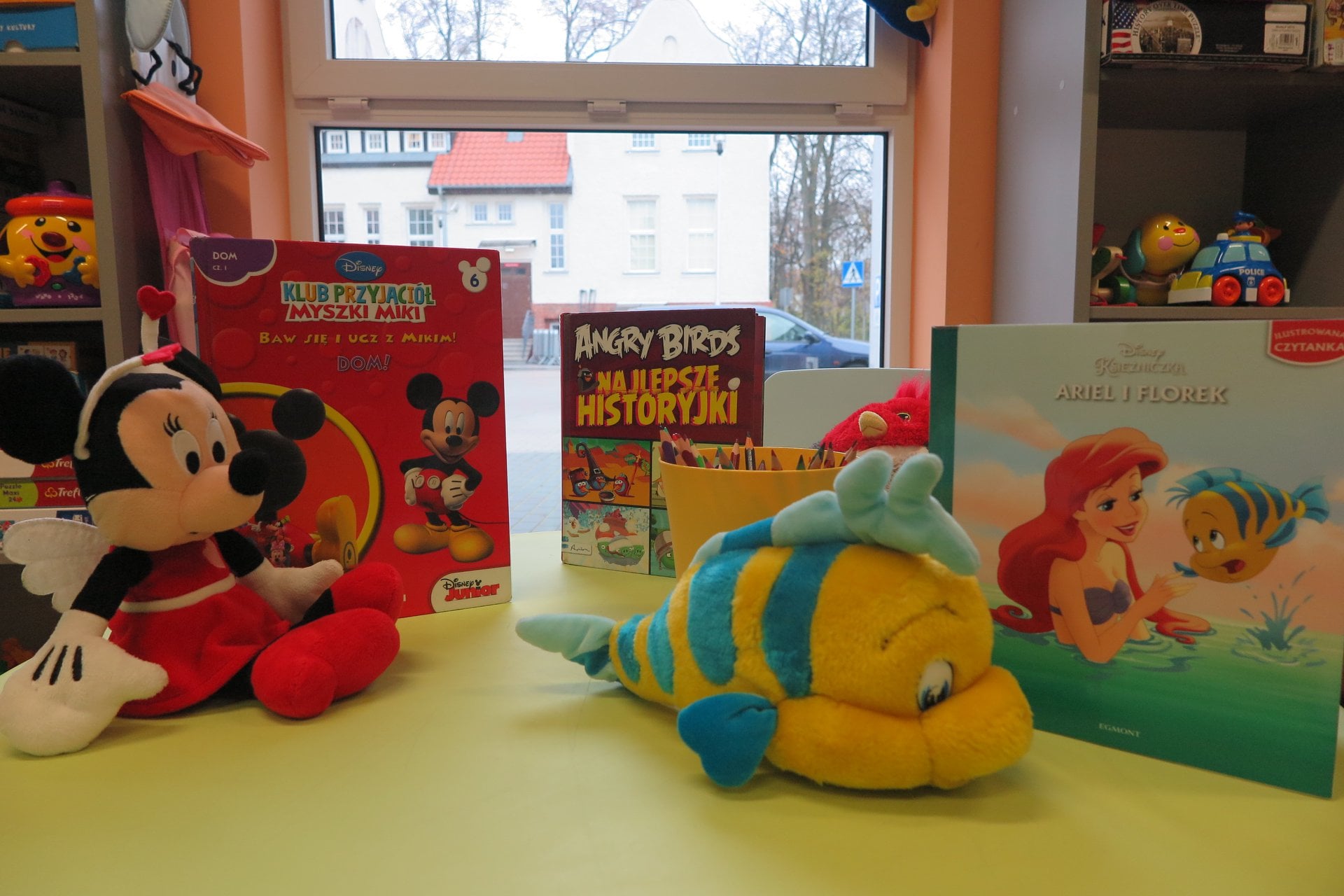 Na stoliku książki dla dzieci o tytule ''Klub przyjaciół Myszki Miki'', '''Angry Birds'', ''Księżniczka Ariel i Florek'' obok pluszowa maskotka myszka miki i maskotka rybka florek.