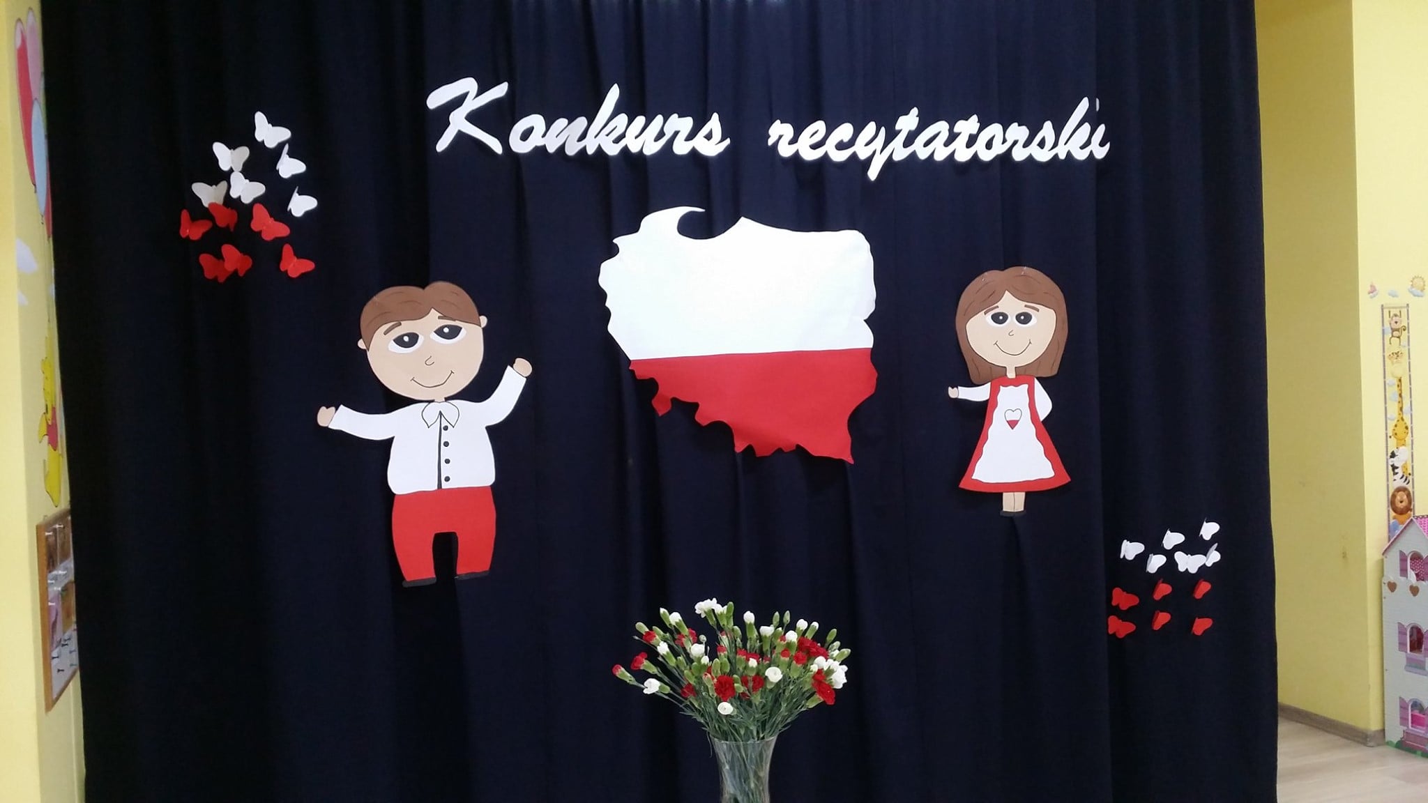 Na niebieskiej zasłonie napis wykonany z papieru pt. ''Konkurs recytatorski'', mapa Polski i dwie postacie chłopiec i dziewczynka ubrani w kolory biało-czerwone. 
