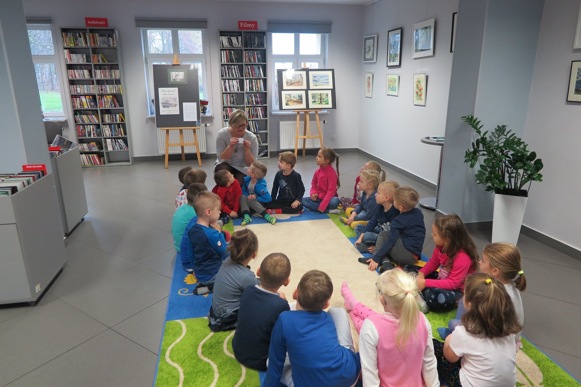 Wnętrze biblioteki, mediateka. Na kolorowym dywanie siedzą przedszkolaki ''Motylki'' z przedszkola nr 5 w Działdowie. Bibliotekarka Dorota Kusik prowadzi warsztaty z programowania. 