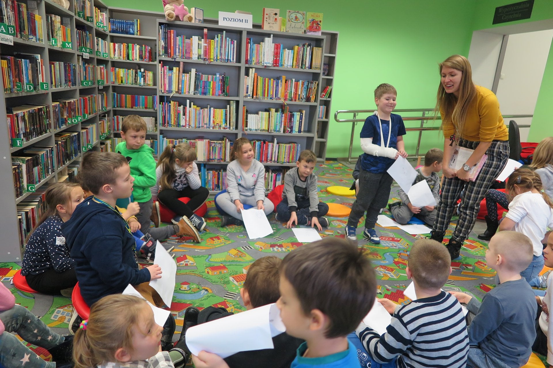 Wnętrze biblioteki. Dzieci z klasy Ib ze SP nr 2 w Działdowie siedzą w kole i trzymają białe kartki. Zajęcia prowadzi bibliotekarka Aleksandra Cybulska. 