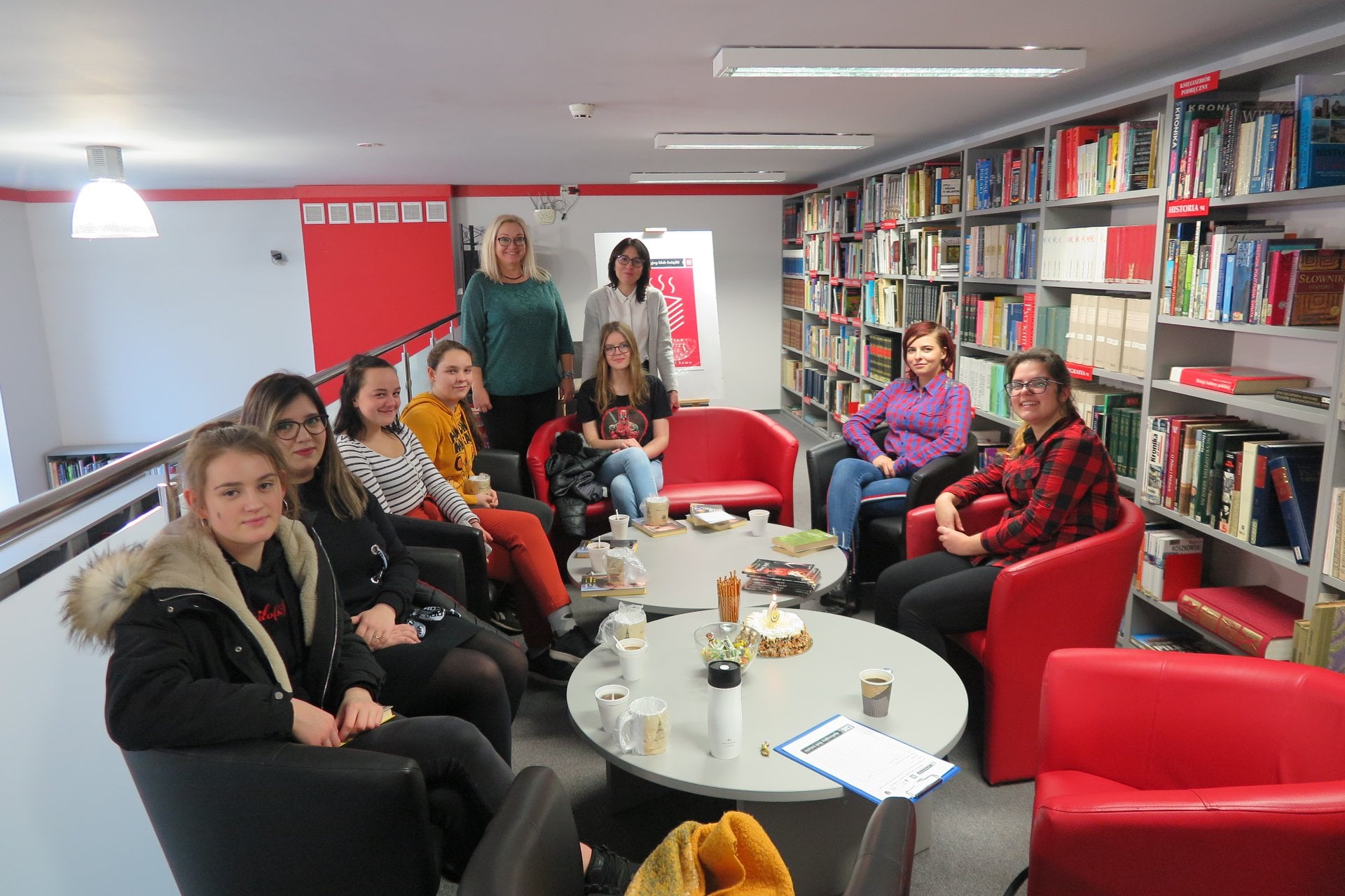 Wnętrze biblioteki. Na kanapach siedzą członkowie klubu DKK ZS Malinowo obok nich stoi pani dyrektor MBP Justyna Lytvyn oraz bibliotekarka ze Szkoły z Malinowa.