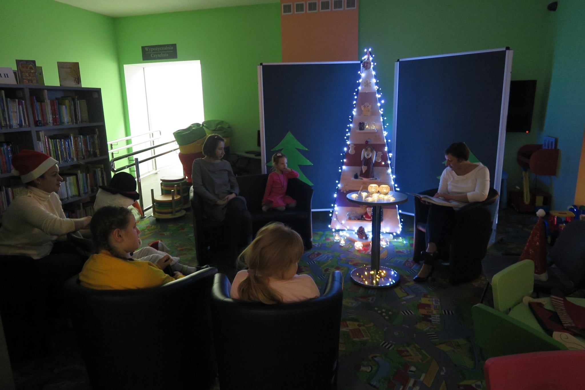 Wnętrze biblioteki, w oddziale dla dzieci spotkała się grupa dzieci żeby czytać świąteczne opowieści oraz śpiewać kolędy. Zajęcia prowadzi bibliotekarka Ania Wiśniewska.