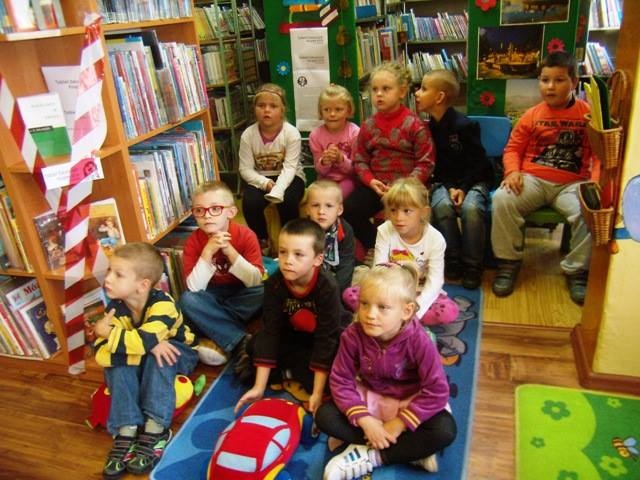 Między regałami z książkami siedzi grupa przedszkolaków biorą udział w  lekcji bibliotecznej.