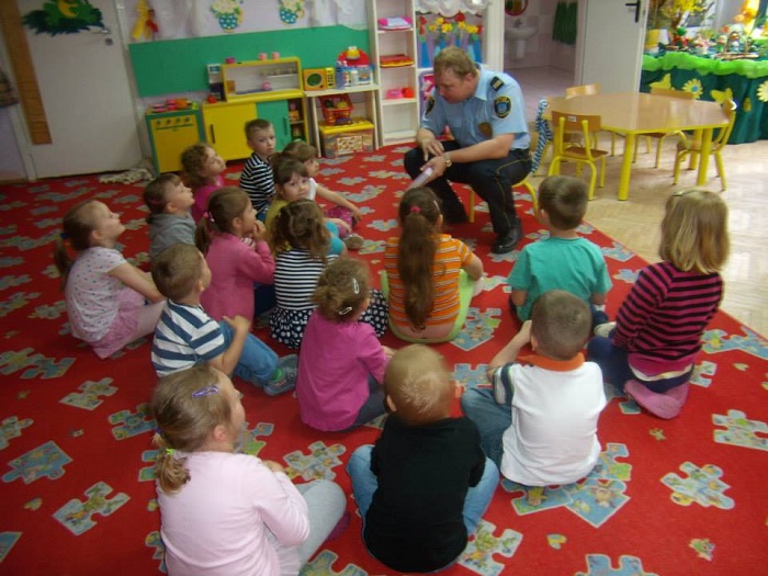 Wnętrze przedszkola, grupa dzieci siedzi na czerwonym dywanie, pan Paweł Buńkowski  ze Straży Miejskiej opowiada dzieciom o przepisach ruchu drogowego oraz zachowania podczas spacerów ze zwierzętami. 