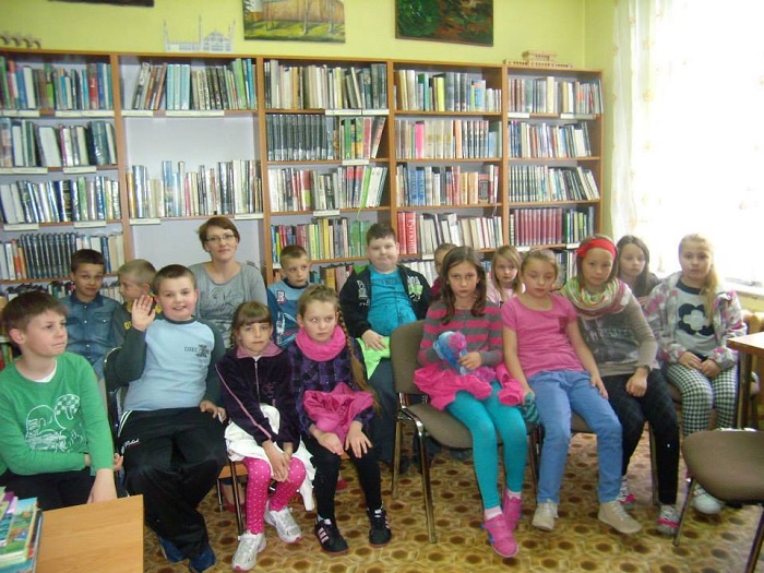Wnętrze biblioteki, na krzesełkach siedzą dzieci biorą udział w lekcji bibliotecznej na temat książki, za nimi regał z książkami.