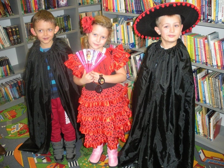 Przy regale z książkami stoi dziewczynka w hiszpańskiej sukience oraz dwóch chłopców w przebraniu Zorro.