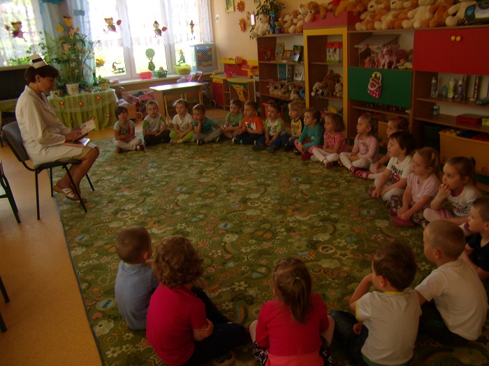 Wnętrze  przedszkola, w kole siedzą dzieci w tle znajdują się zabawki, na krzesełku siedzi Beata Ostrowska z Samodzielnego Publicznego Zakładu Opieki Zdrowotnej w Działdowie czyta dzieciom bajkę.