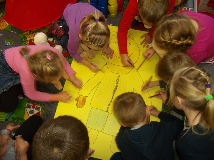 Grupa dzieci koloruje misia narysowanego na żółtym dużym papierze.